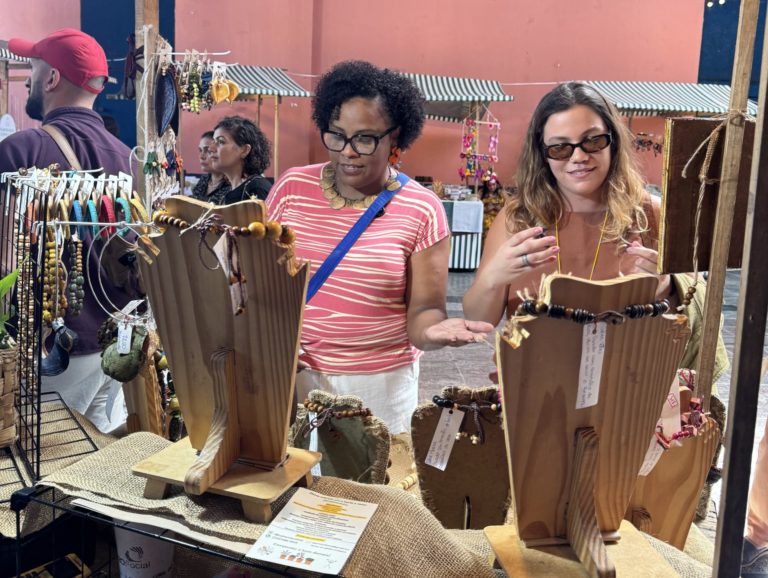 Foto colorida de Márcia Ferreira atendendo uma cliente. Márcia aparece à esquerda, é uma mulher preta com cabelo afro e usa camiseta listrada em branco e vermelho. Ao lado dela, aparece uma mulher branca e loira, que usa óculos escuros e uma blusa marrom