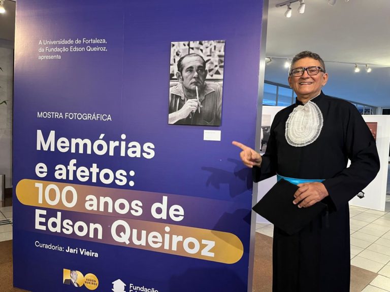 Foto colorida de Joaquim Honorio, vestido com toga, ao lado de banner que diz "Memórias e afetos: 100 anos de Edson Queiroz". Ele é um homem branco, de cabelos grisalhos curtos e usa óculos