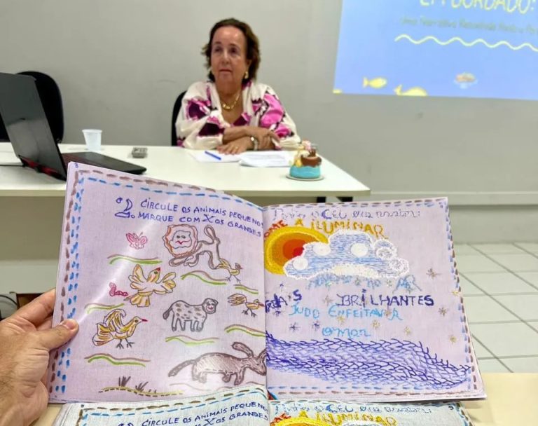Foto colorida que mostra o livro bordado feito por Marivan Ferraro em seu TCC do ensino superior. Na imagem, Marivan aparece ao fundo, sentada. Ela é uma mulher branca, com cabelo preto curto e usa uma blusa florida.