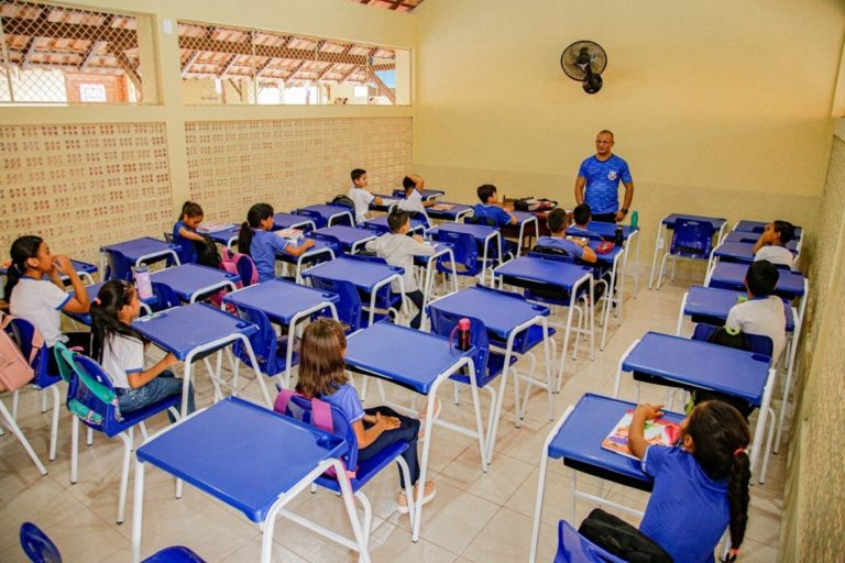 Foto colorida de sala de aula em Santarém. Na imagem, aparece um professor de pé no canto direito da imagem, ele é um homem negro e usa uma camiseta azul. Crianças aparecem sentadas em cadeiras azuis diante de classes azuis.