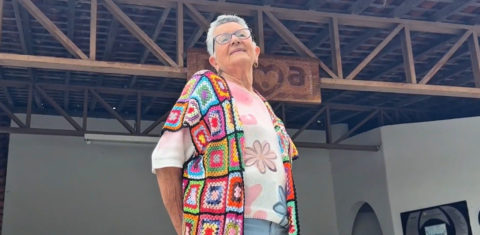 Foto colorida de Socorro Araújo Coutinho. Ela é uma mulher idosa, branca, com cabelo curto branco e usa óculos, uma blusa estampada colorida e sorrindo