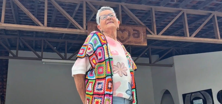 Foto colorida de Socorro Araújo Coutinho. Ela é uma mulher idosa, branca, com cabelo curto branco e usa óculos, uma blusa estampada colorida e sorrindo