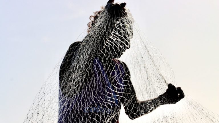 Foto colorida que mostra silhueta de pescador na ilha de Jubim. Na imagem, a pessoa está segurando uma rede de pesca