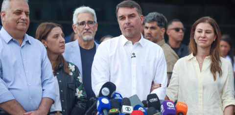 Foto colorida de Flávio Bolsonaro, diante de microfones. Ele está usando camisa branca, ao lado da esposa, Fernanda, que olha para o lado de cabeça baixa, e da economista Daniela Marques