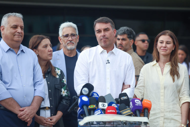Foto colorida de Flávio Bolsonaro, diante de microfones. Ele está usando camisa branca, ao lado da esposa, Fernanda, que olha para o lado de cabeça baixa, e da economista Daniela Marques
