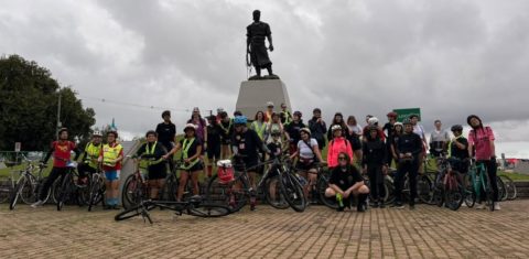 Foto colorida de grupo de mulheres do Pedal das Gurias, reunidas com suas bicicletas em frente à estátua do Laçador, em Porto Alegre