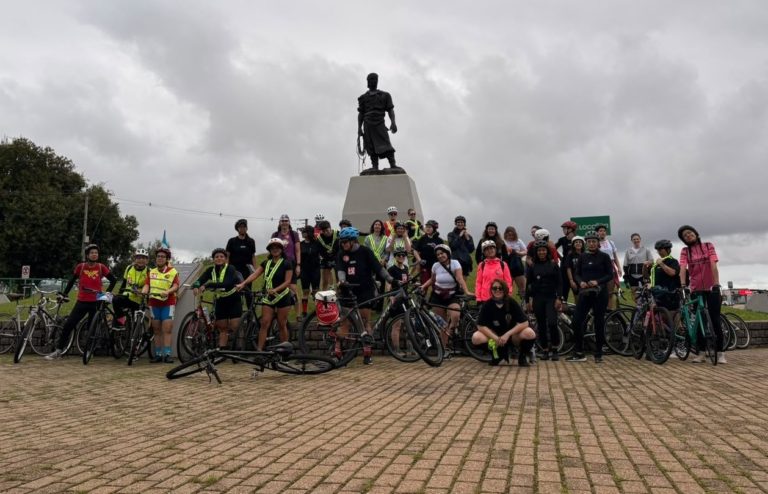 Foto colorida de grupo de mulheres do Pedal das Gurias, reunidas com suas bicicletas em frente à estátua do Laçador, em Porto Alegre