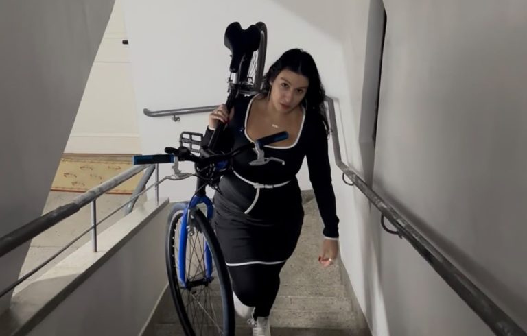 Foto colorida de Tirza subindo escada com sua bicicleta no ombro. Ela é uma mulher branca, com cabelo preto longo e usa um conjunto de blusa e calça preta, com detalhes brancos