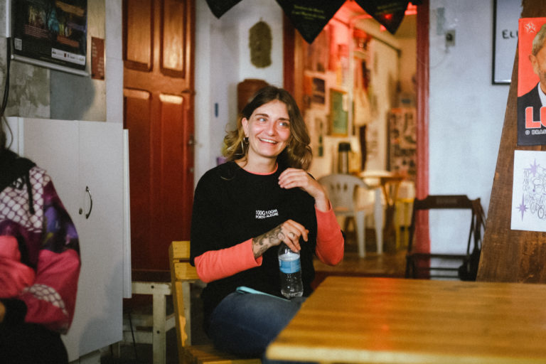 Foto colorida de Paula Moizes. Ela é uma mulher branca, com cabelo loiro e está sorrindo. Ela aparece sentada, usando uma blusa preta e segurando uma garrafa de água