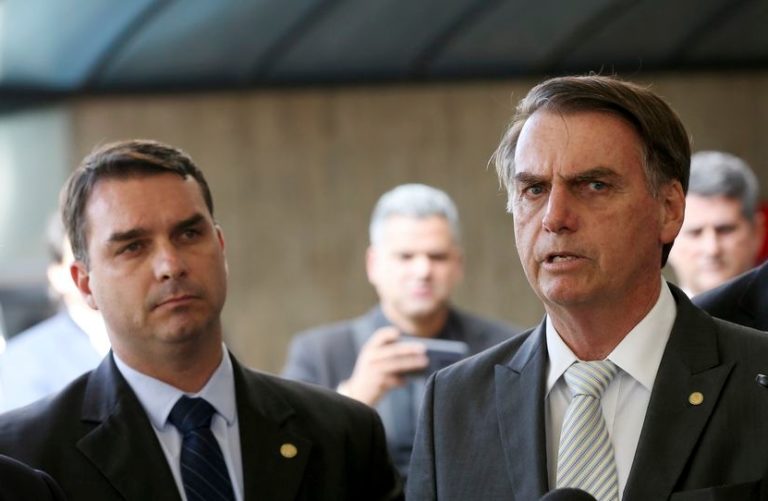 Foto colorida de Flávio (esquerda) e Jair (direita) Bolsonaro. Ambos são homens brancos, usam terno e aparecem com semblante sério.