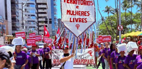 Foto colorida de manifestação do MST em Maceió, com destaque para mulher que segura estandarte onde se lê: territórios e mulheres livres
