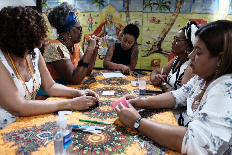 Foto colorida de várias mulheres negras sentadas em volta de uma mesa.