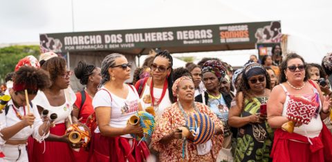 Foto colorida que mostra várias mulheres negras durante marcha das mulheres negras. Elas estão em pé e segurando instrumentos de percussão