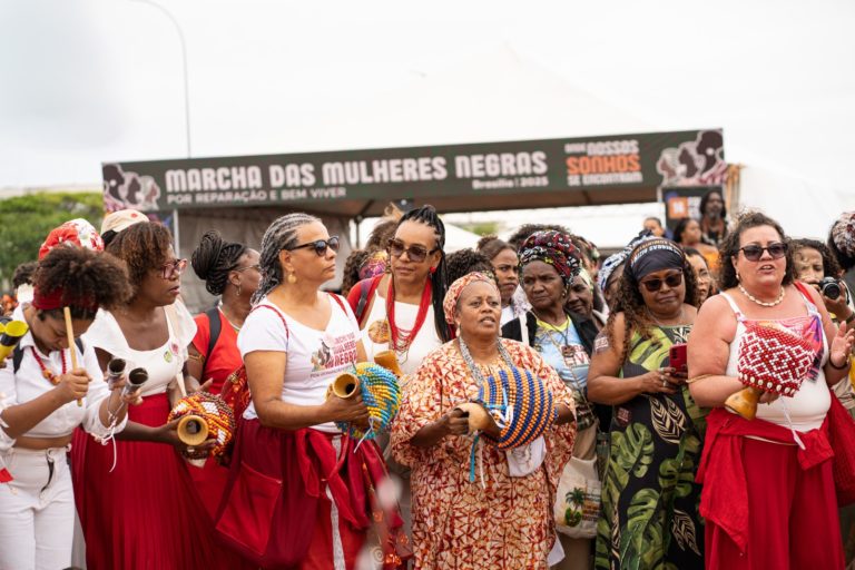 Foto colorida que mostra várias mulheres negras durante marcha das mulheres negras. Elas estão em pé e segurando instrumentos de percussão