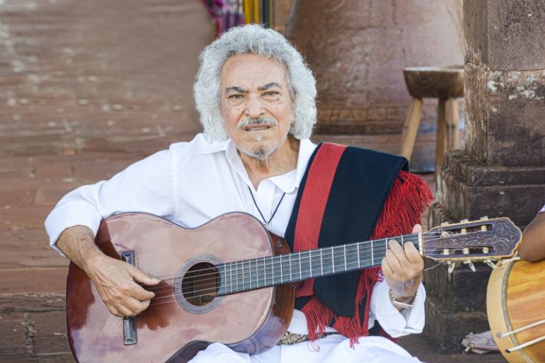 Foto colorida do cantor Pedro Ortaça, homem idoso, branco com cabelo e barba branca e segurando um violão. Ícone da música missioneira
