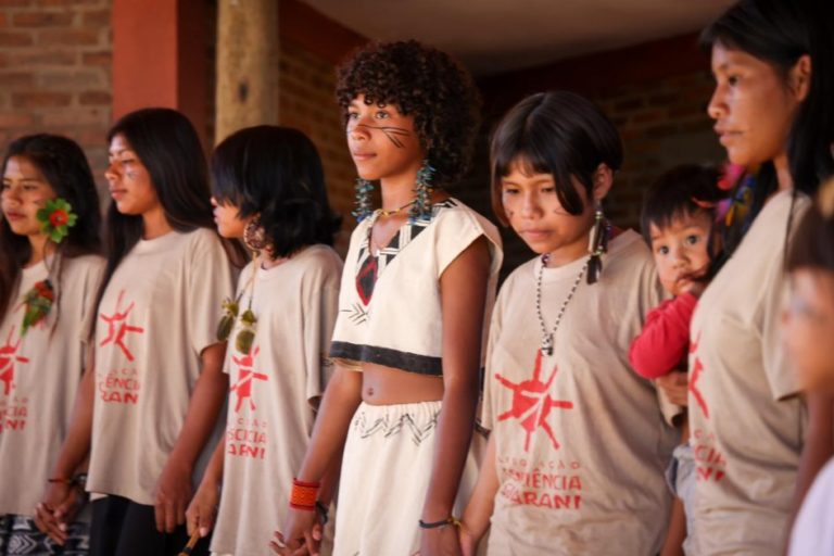 Crianças guarani do coral Jerojy Mbaraete. Elas estão lado-a-lado, de mãos dadas e usam uma camiseta da Associação Consciência Guarani