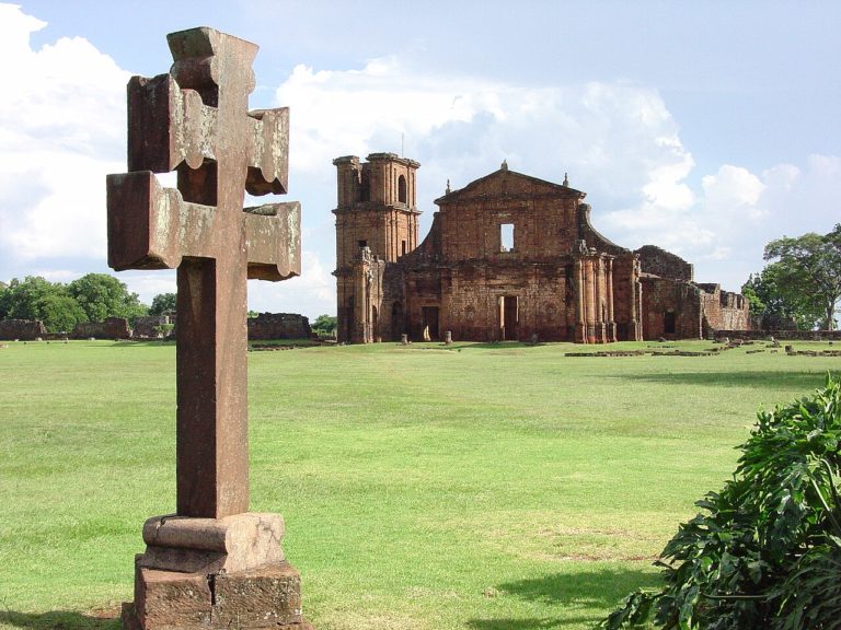Foto colorida da cruz missioneira e, ao fundo, as ruínas de São Miguel das Missões