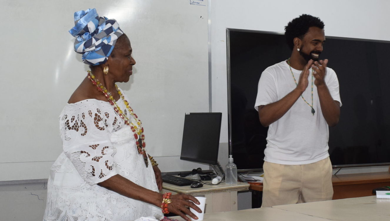 Foto colorida de Mãe Isabel, mulher preta com vestido branco, e Frederico Mendes, homem preto de cabelo e camiseta branca. Ambos aparecem em pé e sorrindo. Frederico está batendo palmas