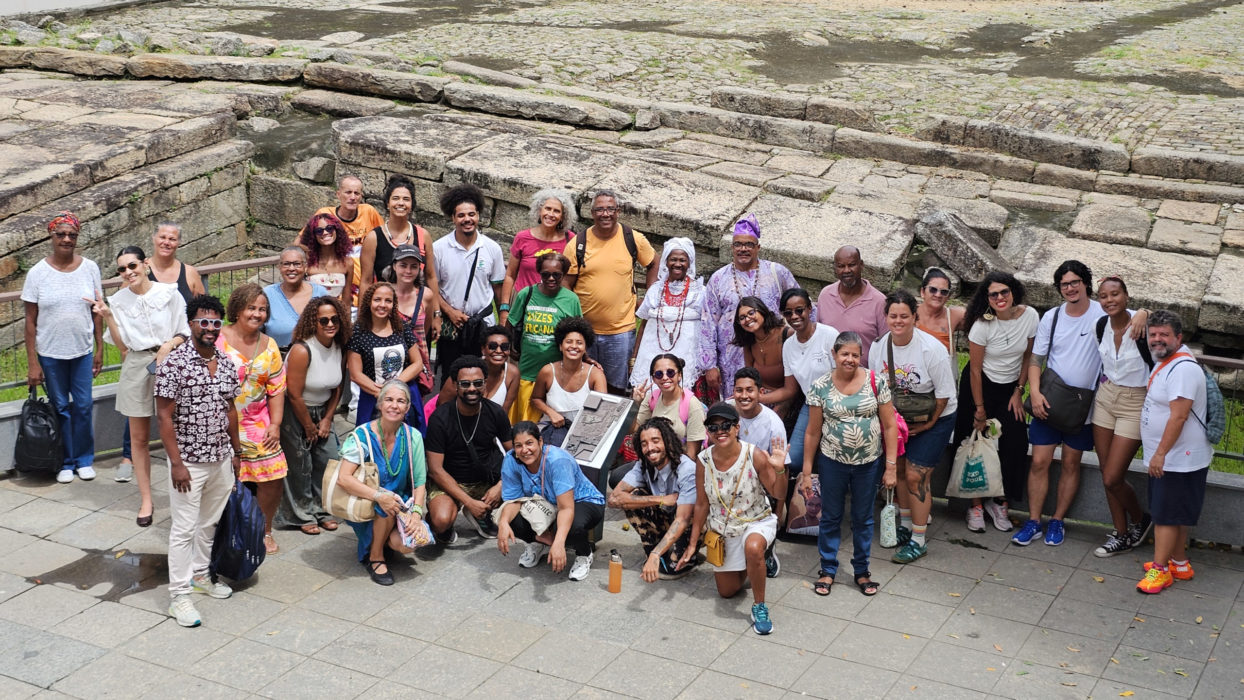 Foto colorida de grupo de pessoas reunidas na Pequena África (RJ). Elas estão de pé e olham para a foto