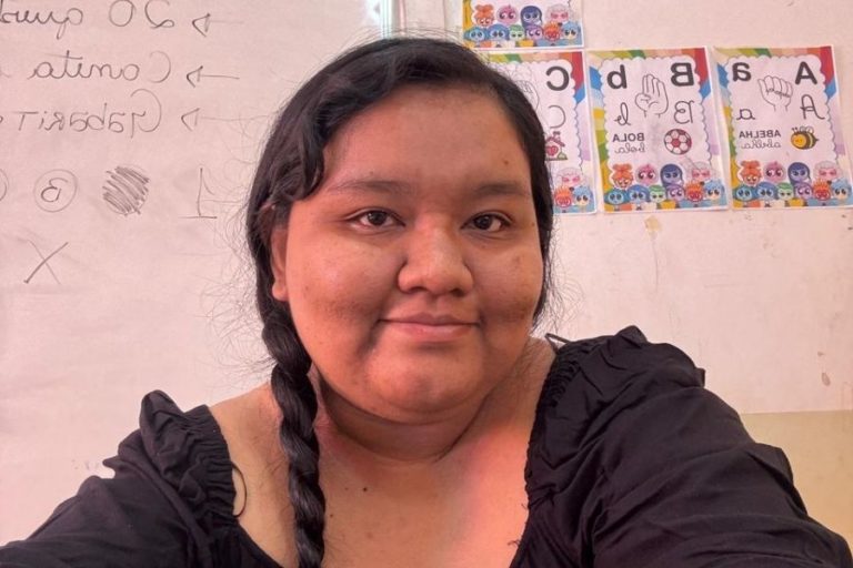 Foto colorida de Glycya Makuxi. Ela é uma indígena de cabelos pretos, que estão enrolados em uma trança. Ela olha para frente. Ao fundo, quadro com desenhos e palavras